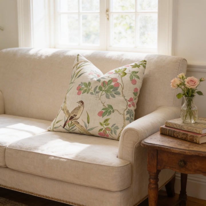 A cozy living room with a beige sofa adorned with a floral pillow, a wooden side table with a vase of flowers, and a stack of books on the table.
