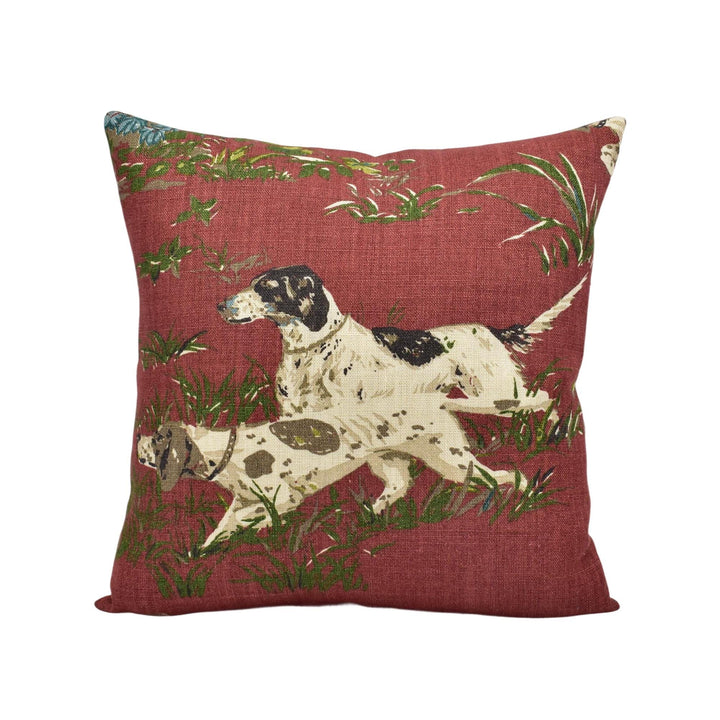 A red throw pillow with a design of two dogs, one black and white and the other brown and white, running through a grassy area with green foliage.