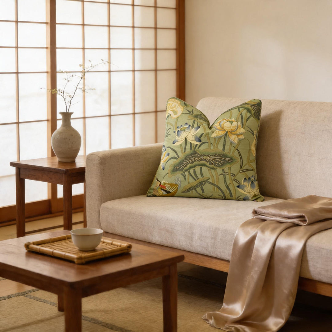 a cozy living room with a beige sofa adorned with a green and yellow floral throw pillow, a wooden coffee table, and a white vase with flowers. The room is illuminated by natural light coming through a window with wooden blinds, creating a warm and inviting atmosphere.