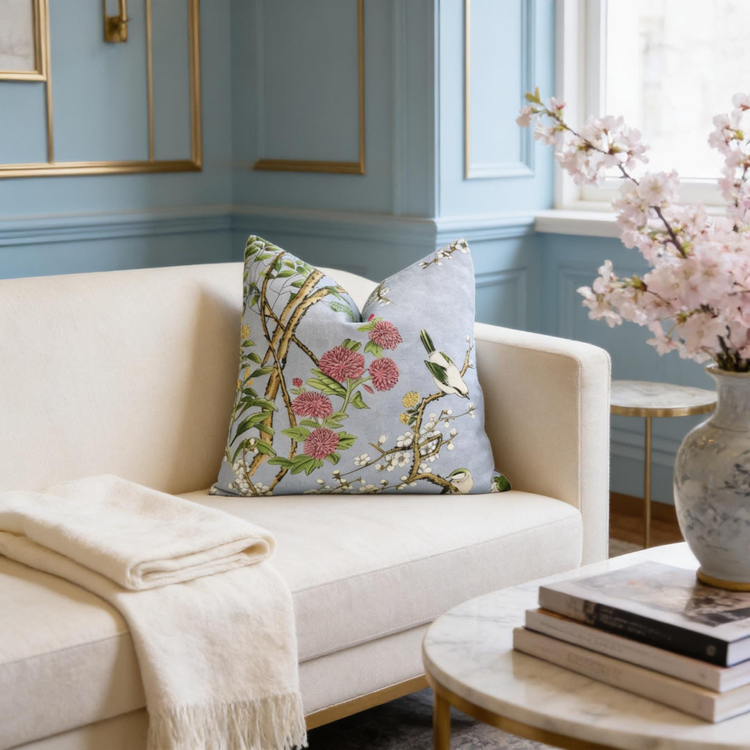 a cozy living room with a white sofa adorned with a decorative pillow featuring a floral design. A white shawl is draped over the sofa, and a vase of pink flowers is placed on a small table nearby. The room has blue walls and gold accents, creating a harmonious and elegant atmosphere.