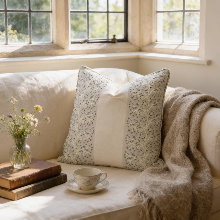 A cozy living room scene with a white sofa, a decorative pillow, a vase of flowers, a stack of books, and a cup of tea on a saucer.