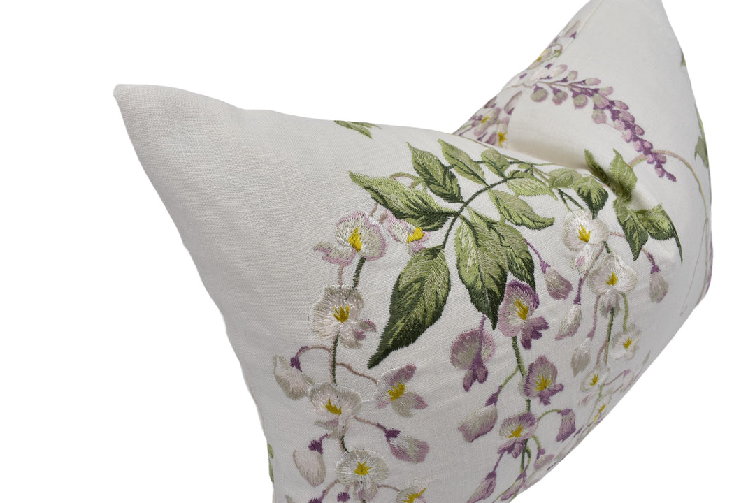 A close-up image of a white pillow with a floral embroidered design featuring purple and white flowers and green leaves.
