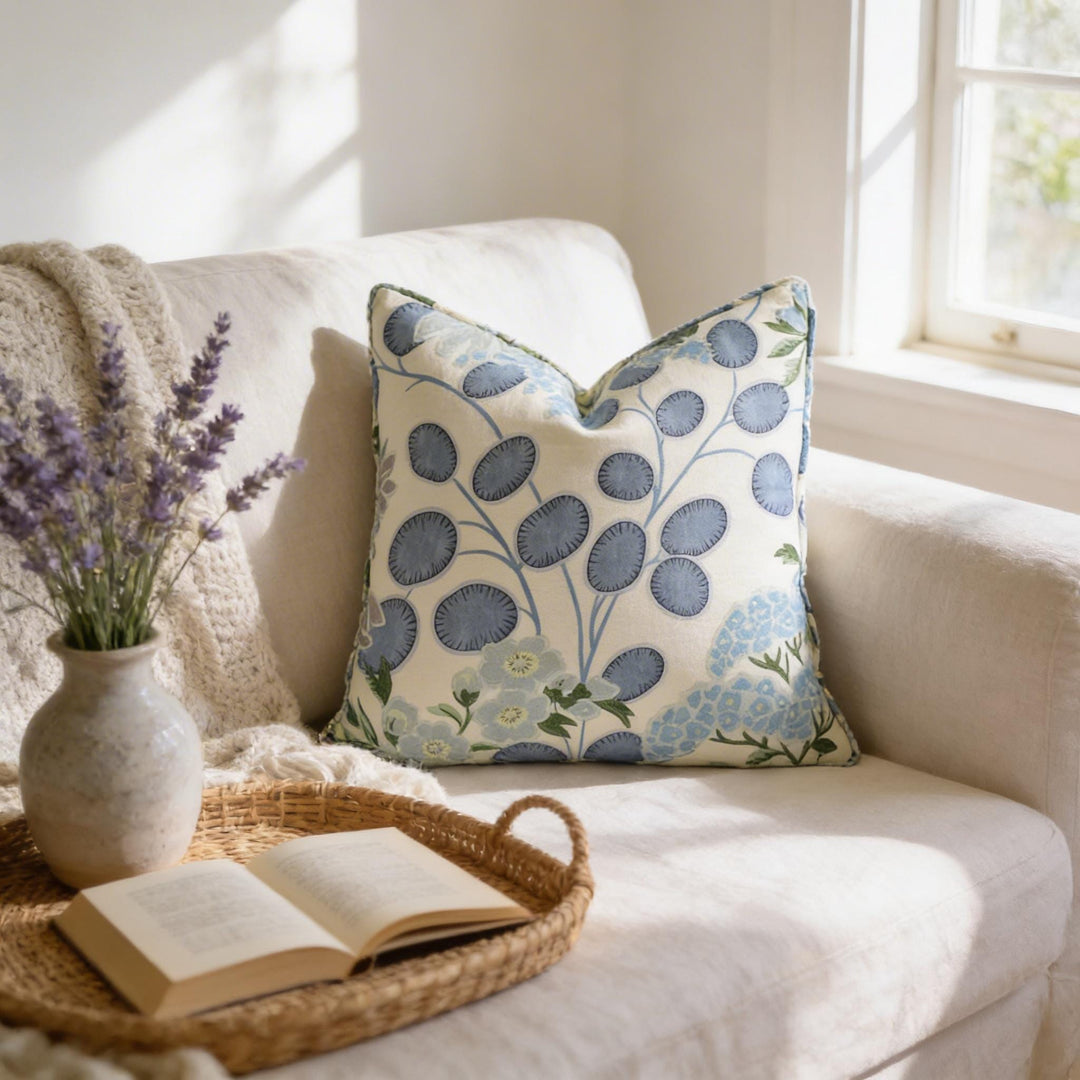 a cozy living room scene with a white sofa, a woven basket with an open book, a vase of lavender flowers, and a decorative pillow with a blue floral pattern.