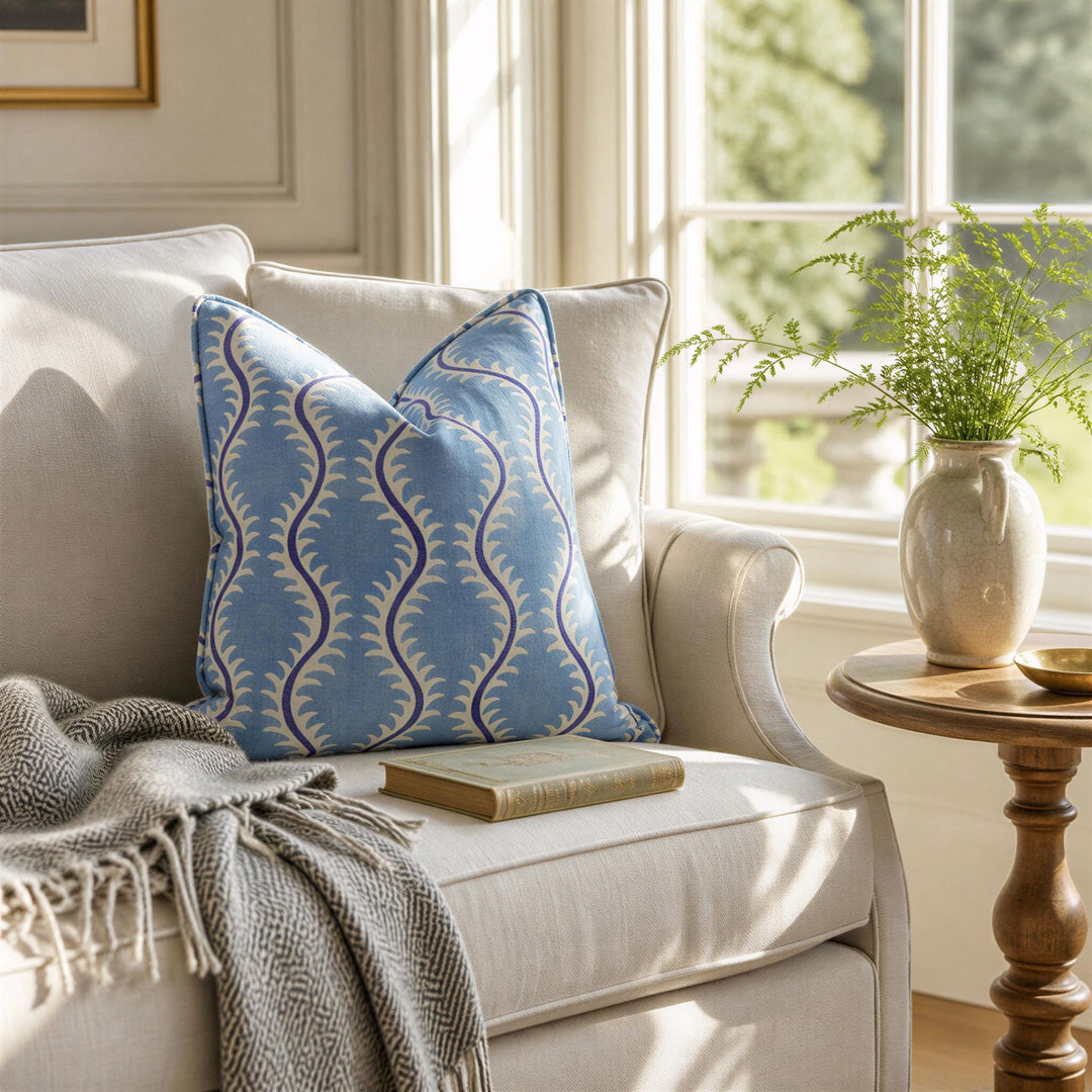 a cozy living room with a white sofa, a blue and white patterned throw blanket, and a wooden side table with a vase of greenery. A book is placed on the sofa, and a window with white curtains lets in natural light, creating a warm and inviting atmosphere.