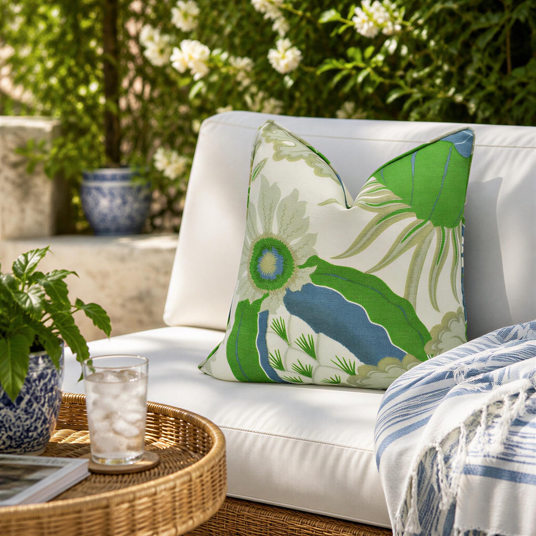 a white outdoor seating area with a green and white floral pillow on a white couch, a wicker table with a glass of water and a plant, and a blue and white striped blanket on the couch.