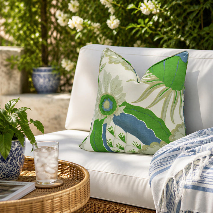 a white outdoor seating area with a green and white floral pillow on a white couch, a wicker table with a glass of water and a plant, and a blue and white striped blanket on the couch.