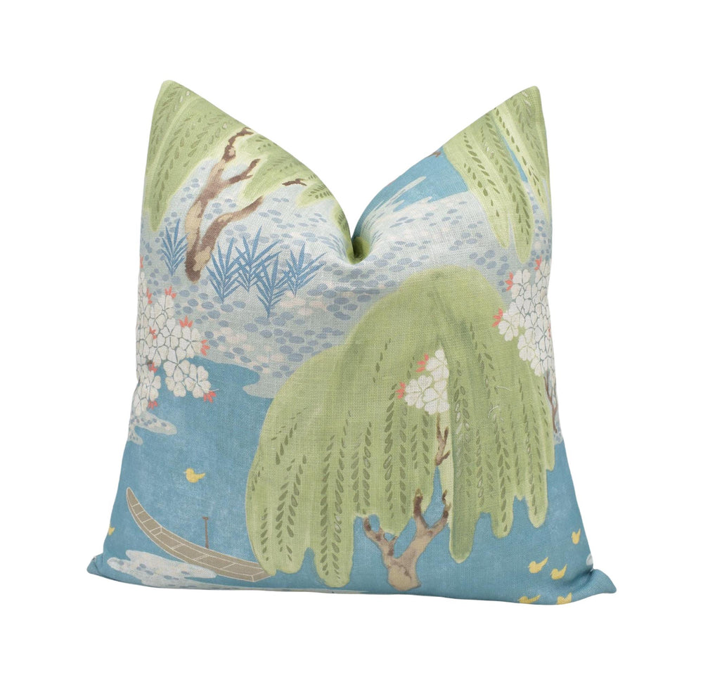 a decorative pillow with a vibrant, nature-inspired design. The pillow has a green and blue color scheme, with a central image depicting a tree with green leaves and a blue sky. Surrounding the tree are various other elements, including a boat, flowers, and birds. The overall design creates a lively and visually appealing scene.