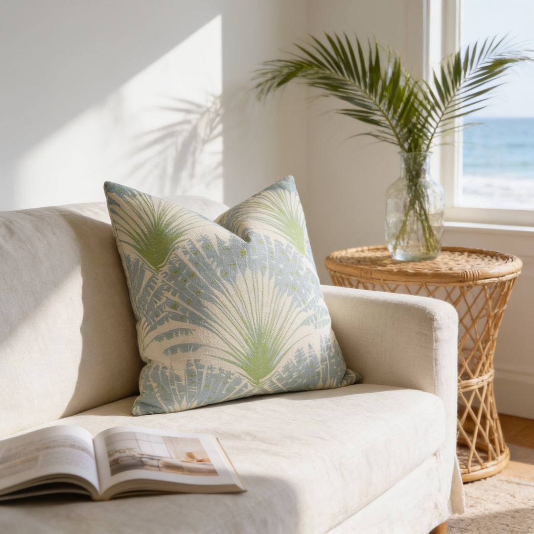 a cozy living room with a white sofa, a decorative pillow, and a wicker side table with a vase of green leaves. An open book is placed on the sofa, and the room has a window overlooking the ocean.
