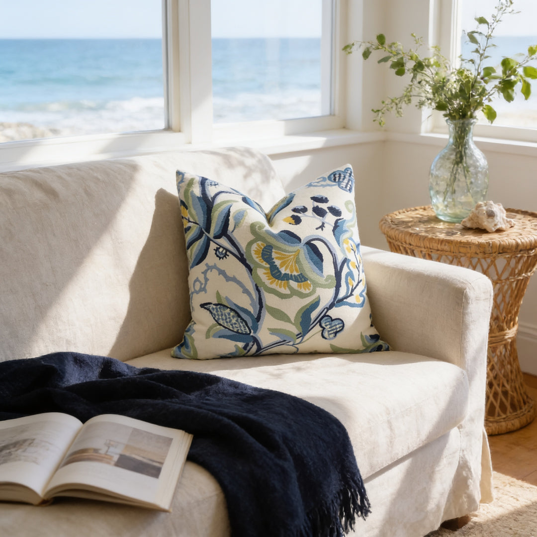 a cozy living room with a beige sofa, a blue and white floral throw pillow, and a woven side table with a vase of greenery. There is an open book on the sofa, and the room has large windows overlooking the ocean, creating a serene and relaxing atmosphere.