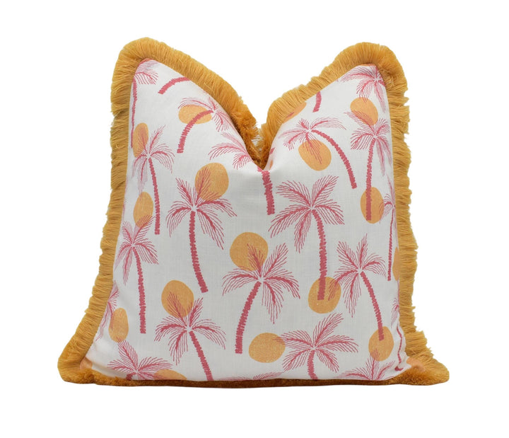 A white pillow with a pattern of pink and orange palm trees printed on it, along with yellow sun-like shapes. The pillow has a fringe border around the edges.