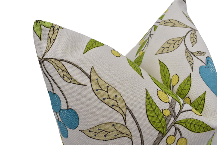 a close-up view of a decorative pillow with a floral pattern. The pillow has a white background and features a mix of green and blue leaves and flowers, along with some blue berries. The design is intricate and visually appealing.