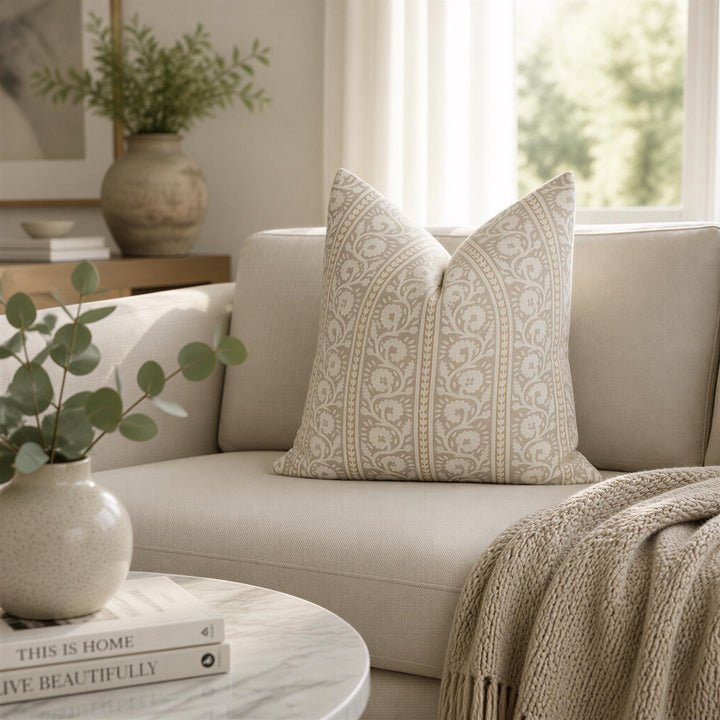 a cozy living room with a beige sofa, a round marble coffee table, and a vase of greenery. The sofa is adorned with a decorative pillow featuring a floral pattern. The coffee table holds a stack of books and a small vase with greenery. The room is illuminated by natural light coming through a window, creating a warm and inviting atmosphere.