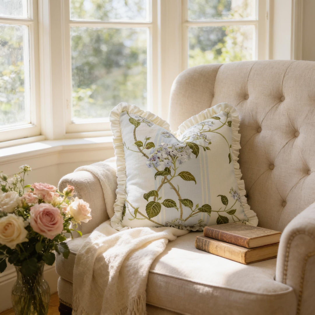 A cozy corner of a room with a tufted beige sofa, a floral throw pillow, and a vase of pink roses. A stack of books is also present on the sofa.