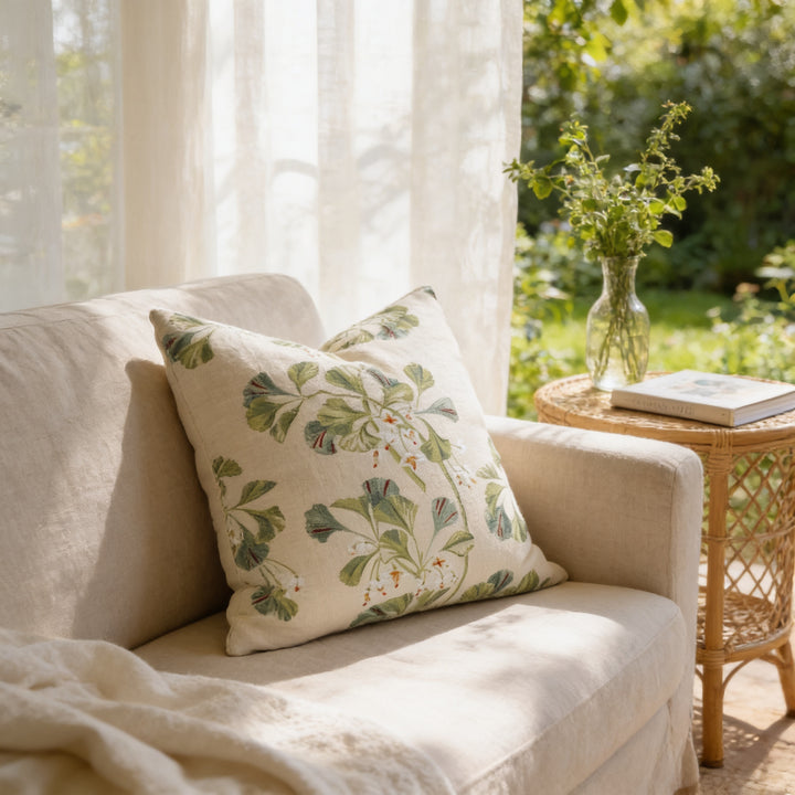 a cozy living room with a beige sofa, a white pillow with a green floral pattern, and a small table with a vase of greenery. The room is illuminated by natural light coming through a window with sheer curtains, creating a serene and inviting atmosphere.