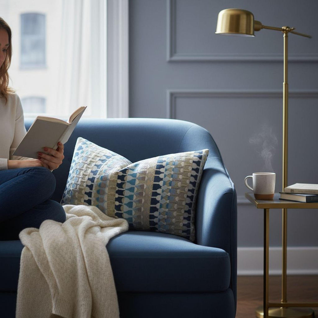A woman is sitting on a blue armchair, reading a book, with a cup of coffee and a lamp nearby.