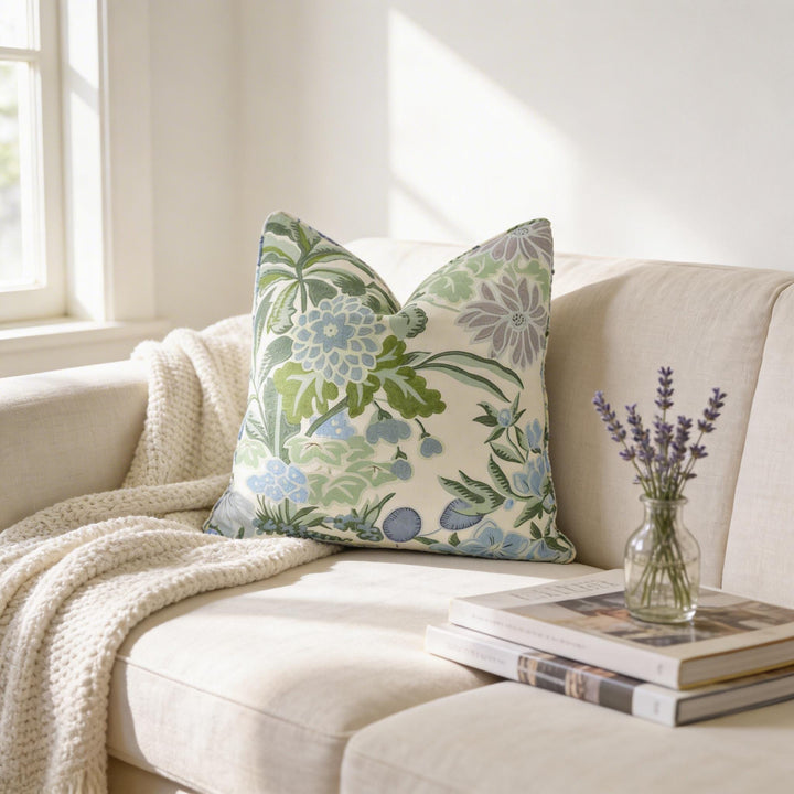 a cozy living room with a beige sofa adorned with a green and white floral throw pillow. A stack of books is placed on the armrest, and a vase of purple flowers is positioned on the right side of the sofa.
