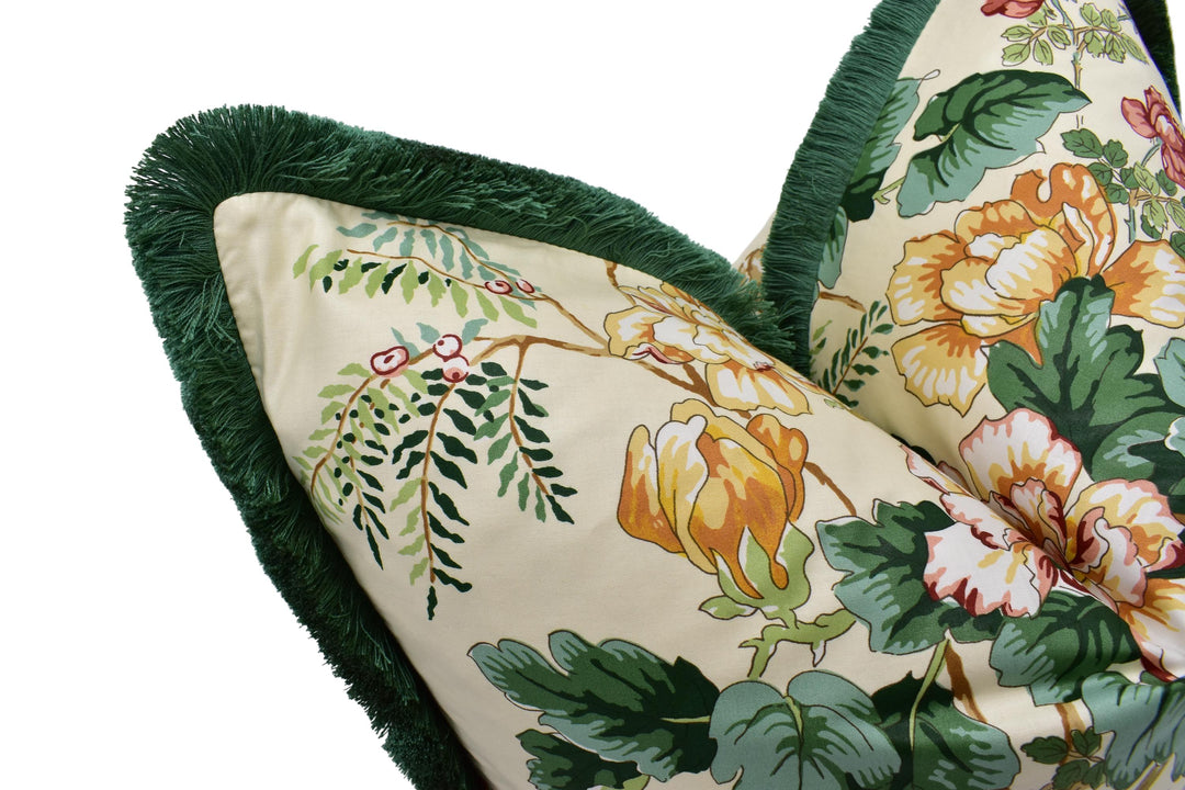 a close-up view of a decorative pillow with a floral pattern. The pillow has a green fringe around the edges and features a vibrant design with yellow and orange flowers, green leaves, and other natural elements.