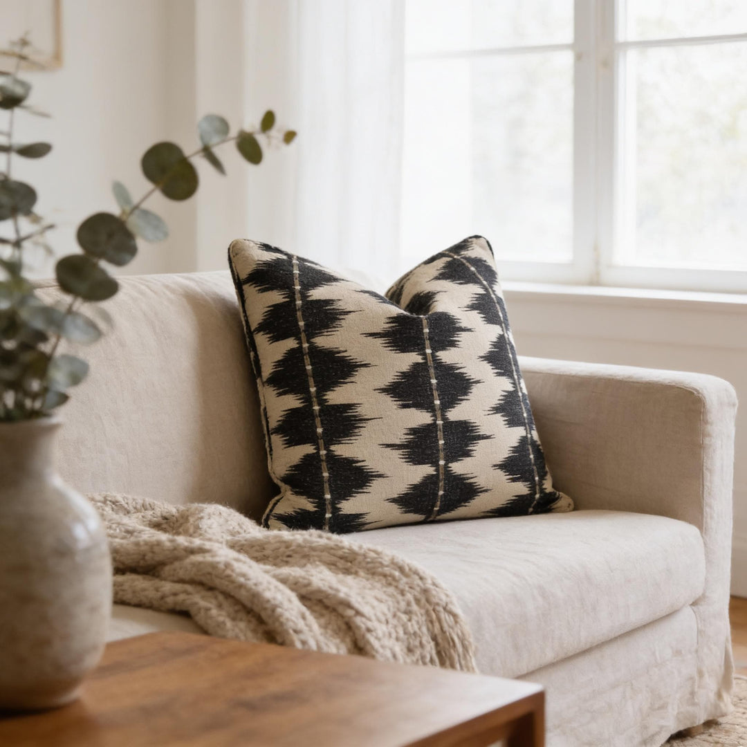 a cozy living room with a beige sofa adorned with a black and white patterned throw pillow, a wooden coffee table, and a vase of greenery.