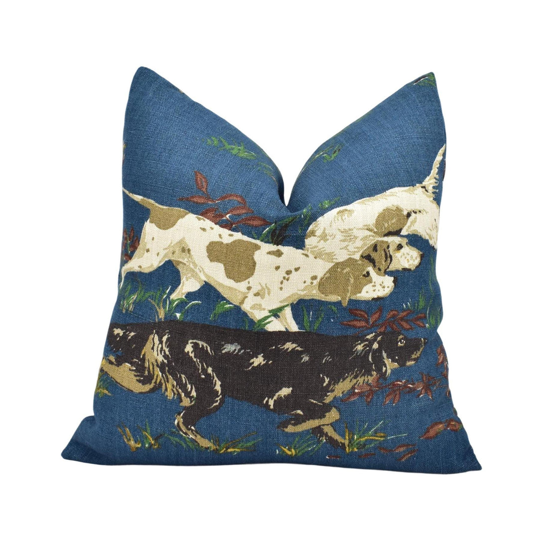 A blue throw pillow with a pattern of two dogs, one white and one brown, running through a grassy field with leaves.