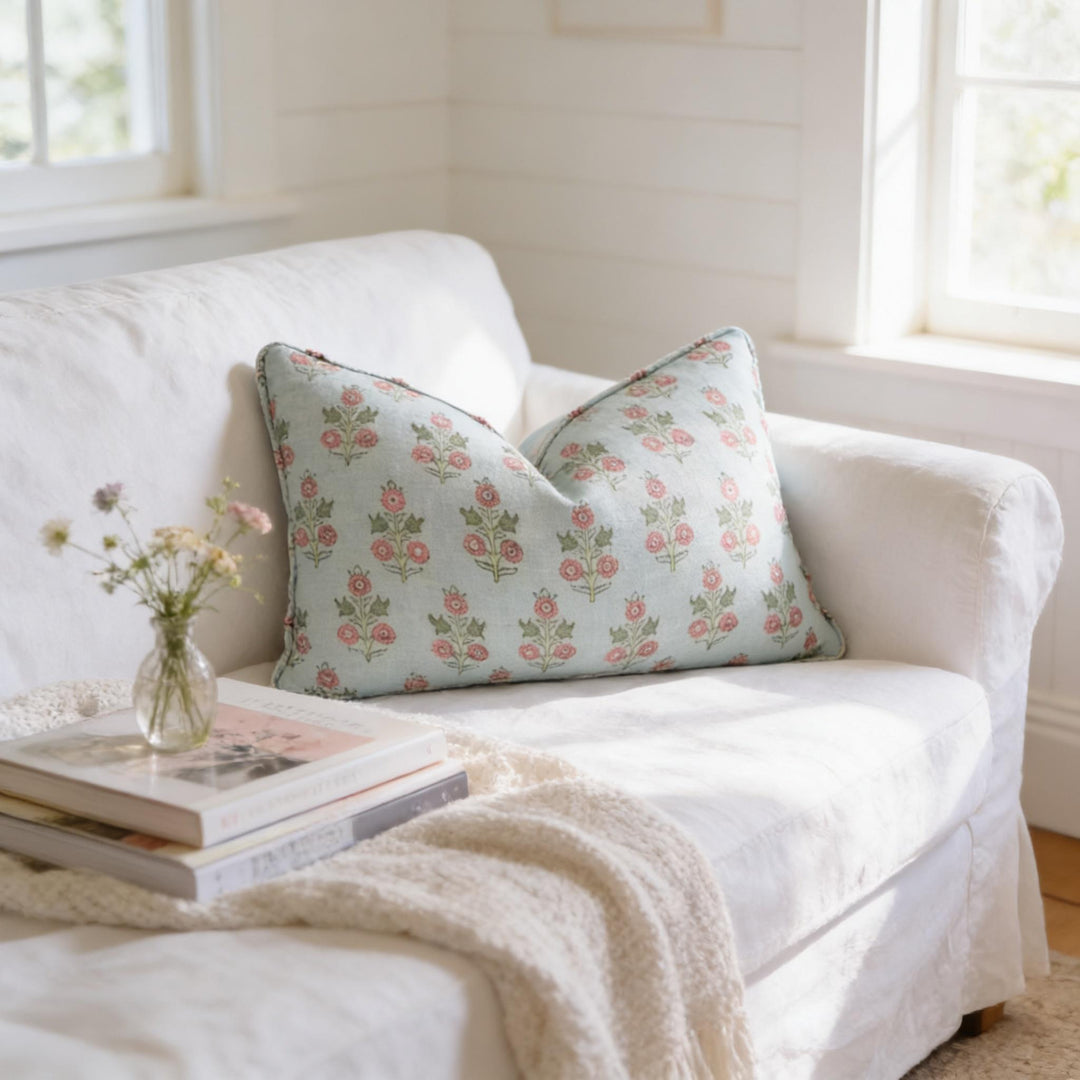 a cozy living room with a white sofa, a floral throw pillow, and a stack of books on the armrest. A vase of flowers is placed on the armrest, adding a touch of nature to the space. The room has a bright and airy atmosphere, with natural light coming in from a window.