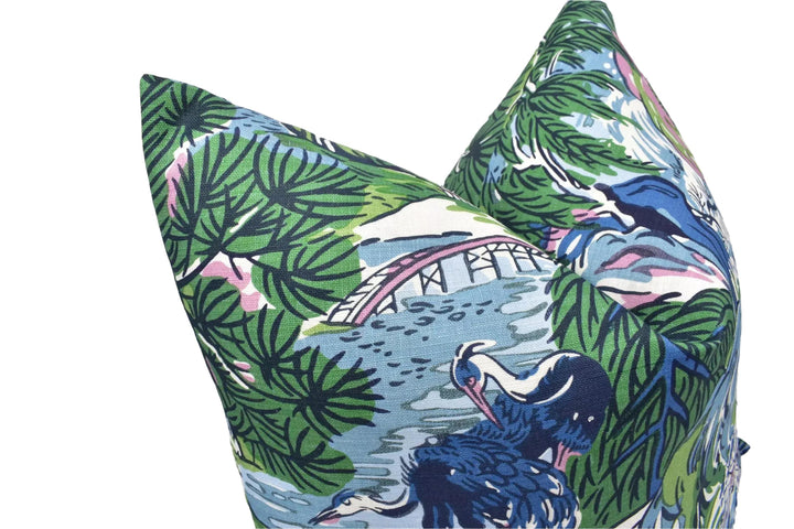a vibrant, colorful pillow with a tropical print design. The pillow has a mix of green, blue, and pink colors, and it depicts various elements such as palm trees, a bridge, and birds. The pillow appears to be a decorative piece, adding a touch of nature and visual interest to the space it is placed in.